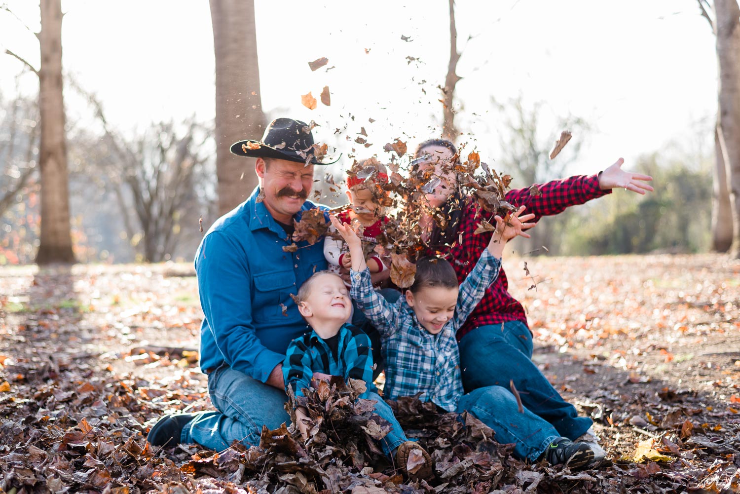 Unposed Fall family photos of family sitting under trees throwing brown leaves up in the air in Delano TN