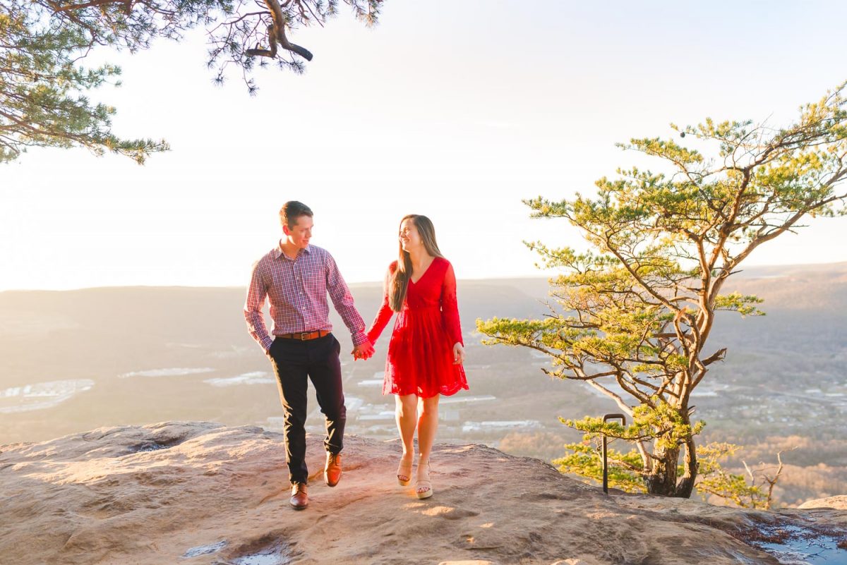 girl in red lace dress and guy in navy pants walking on rock at sunset rock with golden glowing light behind them