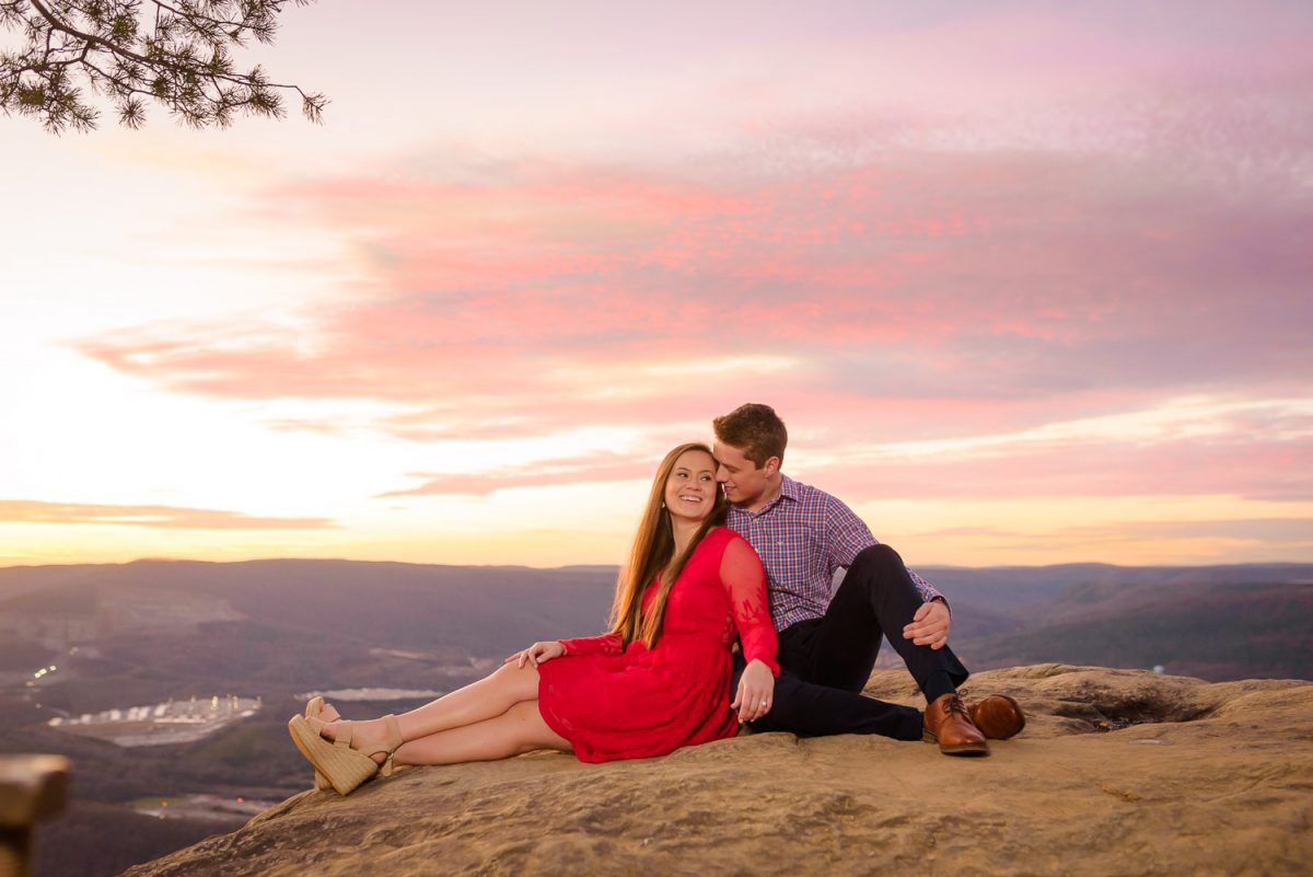 girl in red dress and guy in navy and plaid sitting on rock with glowing pink sky in the background