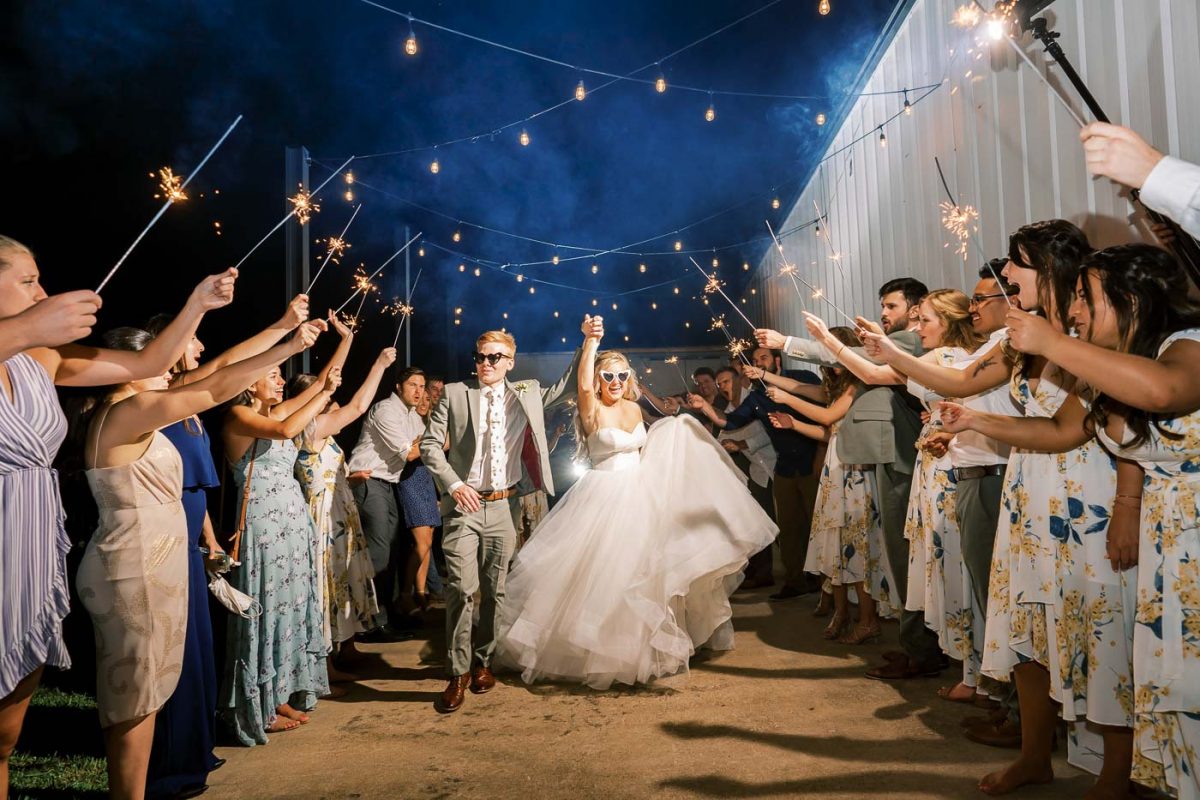 Gwedding guests outside holding sparklers as bride and groom come out wearing sunglasses at night